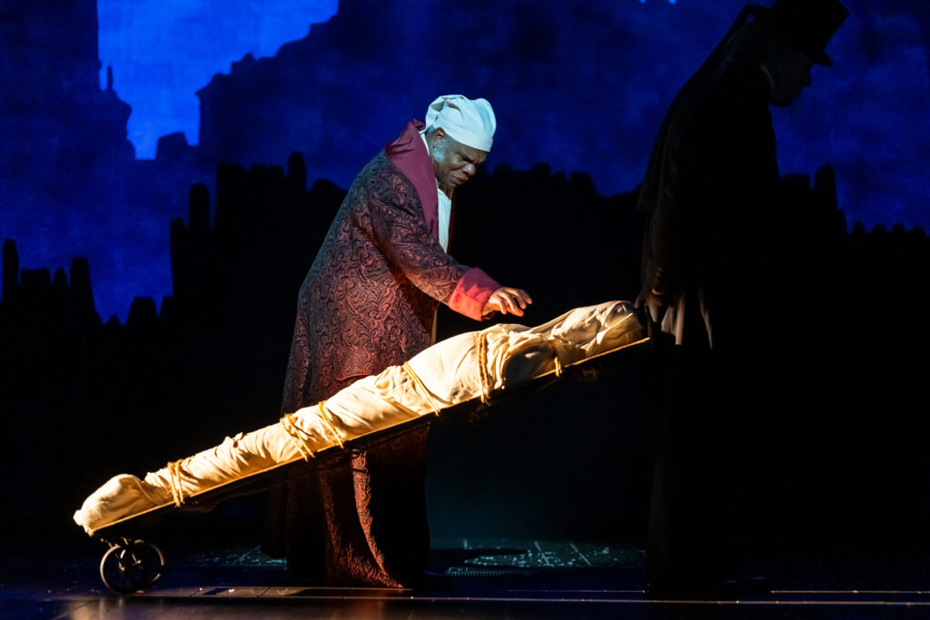 XMASCAROL2024_0201 Social: Craig Wallace as Ebenezer Scrooge in the 2024 Ford’s Theatre production of A Christmas Carol, directed by Michael Baron. Photo by Scott Suchman. E-News/Publications: Craig Wallace as Ebenezer Scrooge in A Christmas Carol (2024). Photo by Scott Suchman. Craig Wallace as Ebenezer Scrooge in the 2024 Ford’s Theatre production of “A Christmas Carol,” adapted by Michael Wilson, directed by Michael Baron. The production plays November 21-December 31, 2024, and features Scenic Design by Lee Savage, Costume Design by Alejo Vietti, Lighting Design by Rui Rita, Original Music and Sound Design by Josh Schmidt, Choreography by Shea Sullivan and Wig Design by Charles G. LaPointe. Photo by Scott Suchman. A Black man wearing a Victorian-era night cap and red house robe with concern on his face reaches out with apprehension as a dead body wrapped in cloth and tied to a dolly is wheeled past him.