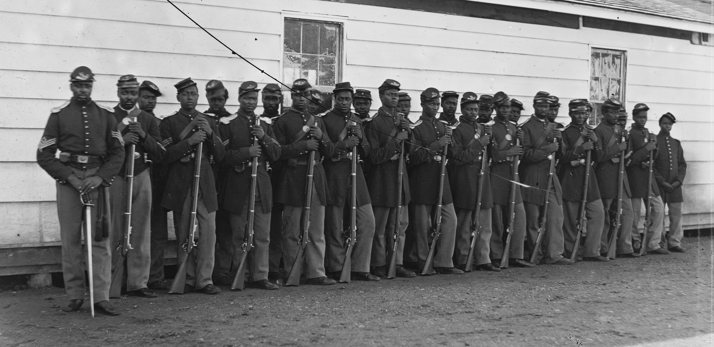 Black and White photograph of a company of African American soldiers dressed in Union Civil War uniforms. They stand in two lines, at attention, posing in front of a barracks.