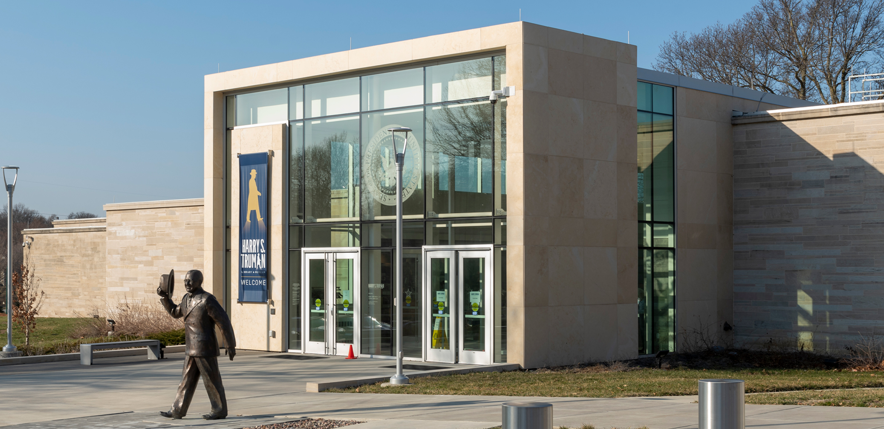 Exterior of a large beige building with ceiling-height windows at the entrance and a bronze statue of a man walking in front.