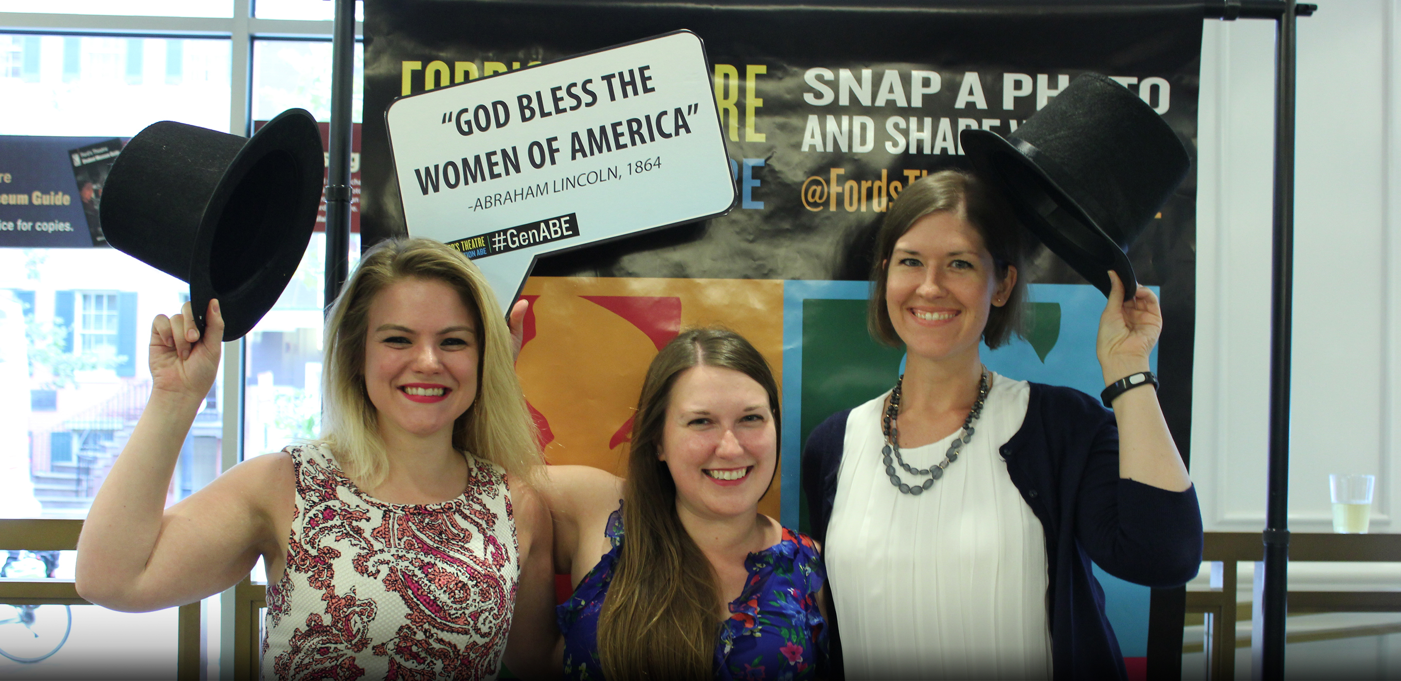 Three woman stand arm in arm in the lobby of Ford's Theatre. Two are holding stovepipe hats. One is holding up a sign that reads "God Bless the Women of America - Abraham Lincoln, 1864."