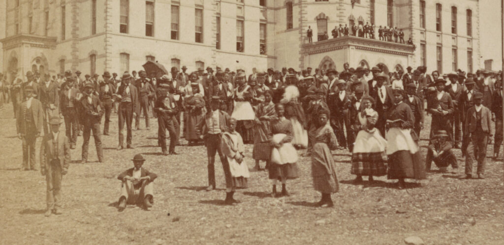 A double image of an enormous and ornate Victorian university building, with a couple of hundred well-dressed, African American men and women arranged in front of it on the lawn for a large group photo. They are students at Howard University around 1867.