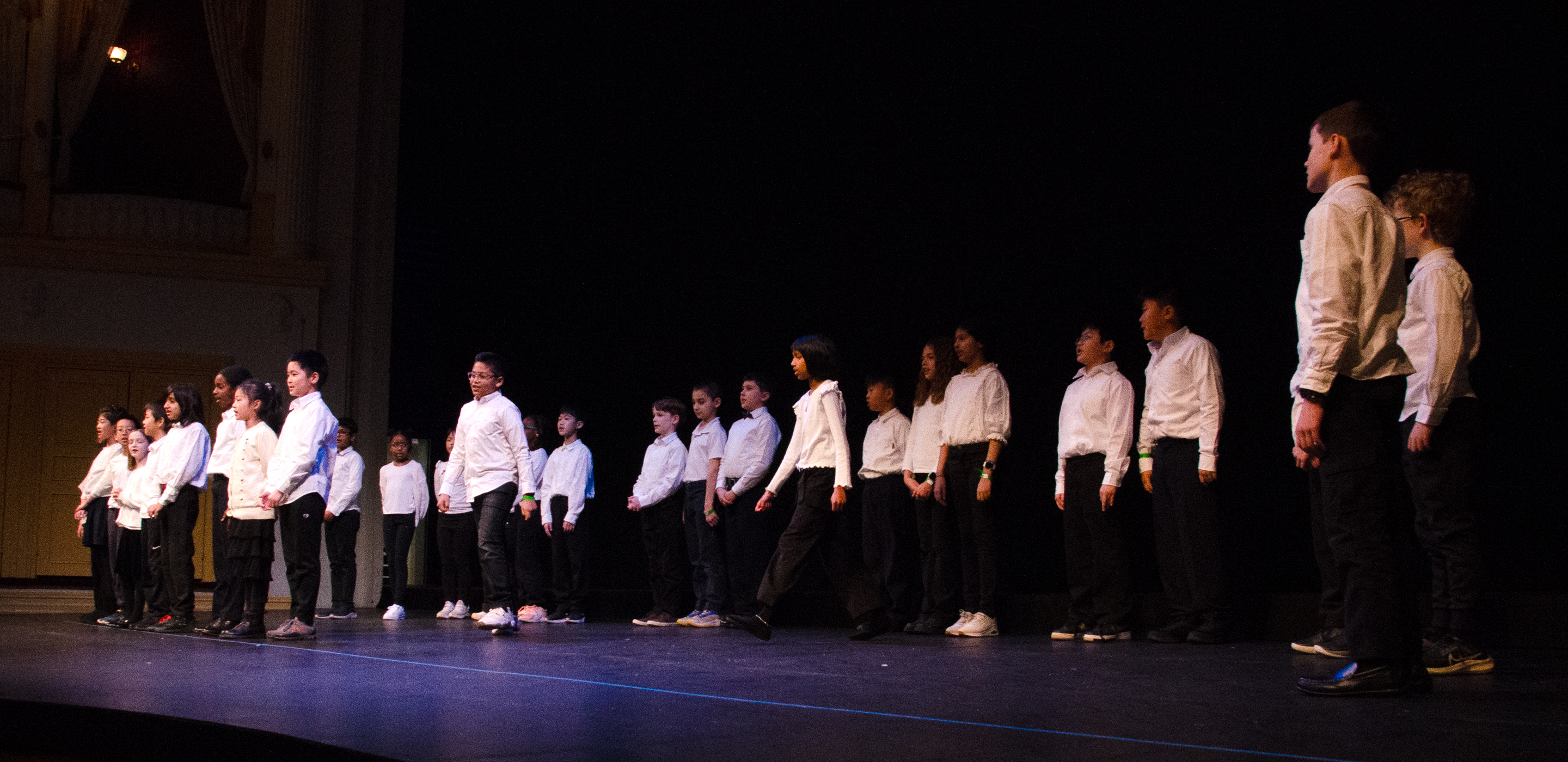 A group of students wearing white shirts and dark pants stand spaced across a dimly lit stage inside Ford’s Theatre. Warm light from the theatre boxes illuminates part of the background as the students face forward some taking a step forward ot being their oratory performance
