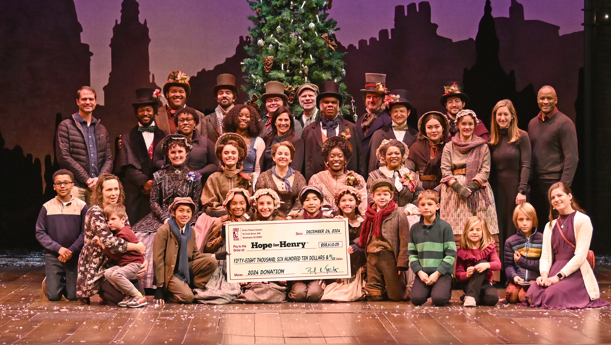 A large group of people, mostly actors in Victorian outfits, pose with a poster-size check on stage with the set and large Christmas Tree behind them.