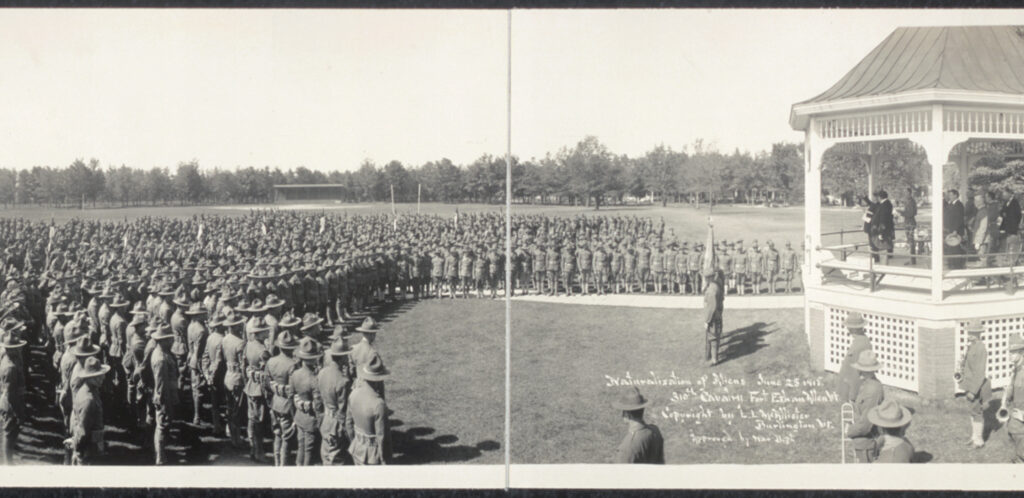 A black and white panorama photograph showing around 100 military servicemembers in formation, facing a gazebo where speakers and officials wearing suits address the crowd.