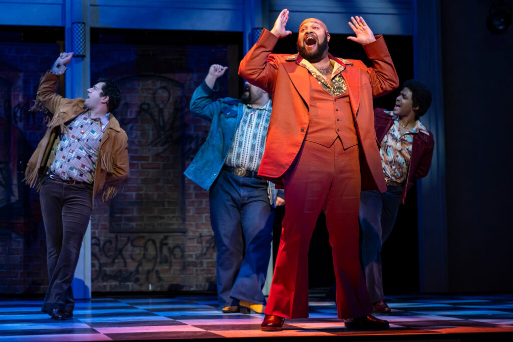A Black man dressed as a 1970s mobster in a bight orange suit, stands center stage and sings in front of three men, dressed in 1970s street clothes appearing as back-up dancers.