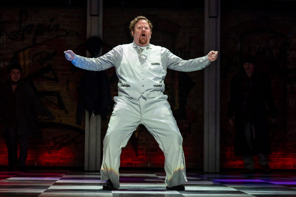 A white man dressed in shiny white dress pants and vest, with a shimmery silver shirt, dances animatedly on stage in a spotlight.