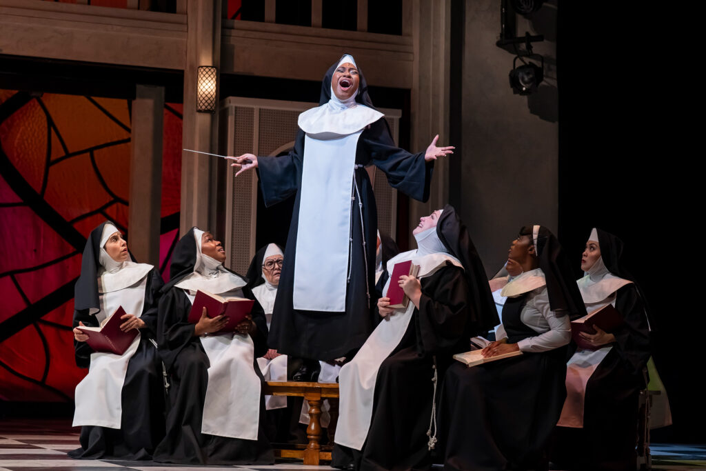 A nun holding a conductor’s wand stands on a bench singing with arms outstretched with a group of nuns sitting around her with expressions that convey surprise, confusion and joy.