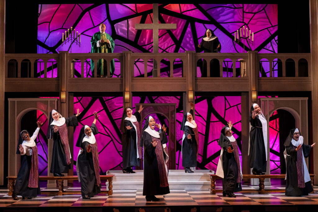A large group of nuns sings and dances animatedly on the stage against a church set with a colorful stained-glass background. Elevated on a balcony behind the nuns, and looking down in prayer are a shocked mother superior and the monsignor.