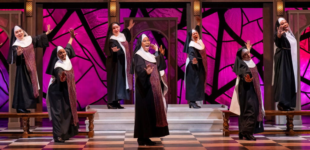 A large group of nuns sings and dances animatedly on the stage against a church set with a colorful stained-glass background. Elevated on a balcony behind the nuns, and looking down in prayer are a shocked mother superior and the monsignor.