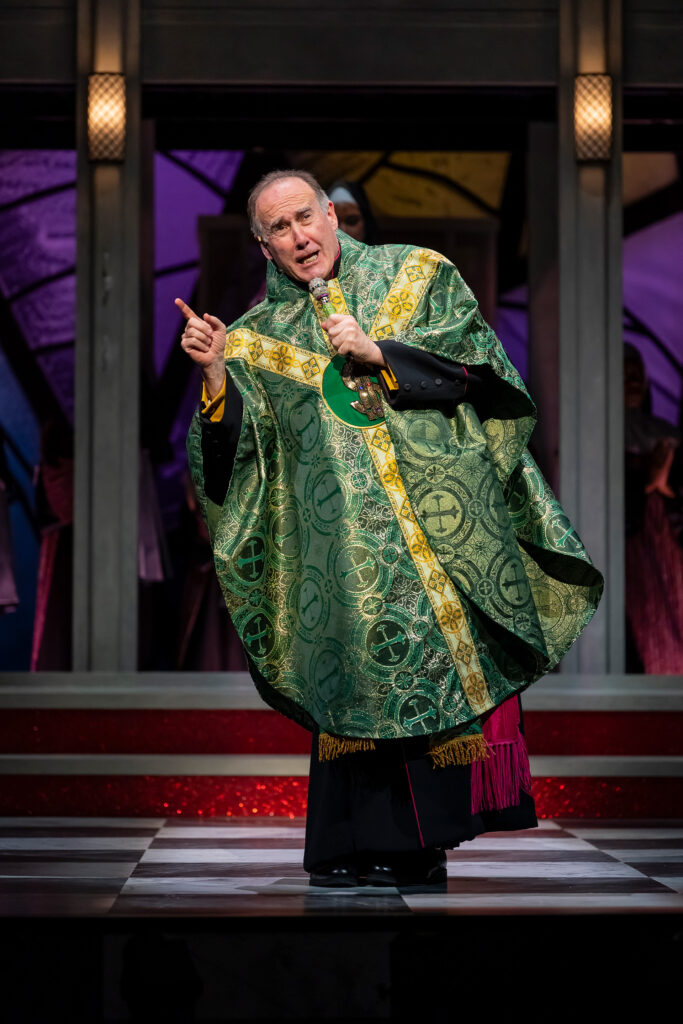 A white man dressed as a catholic priest in a green and gold robe speaks into a microphone he is holding.