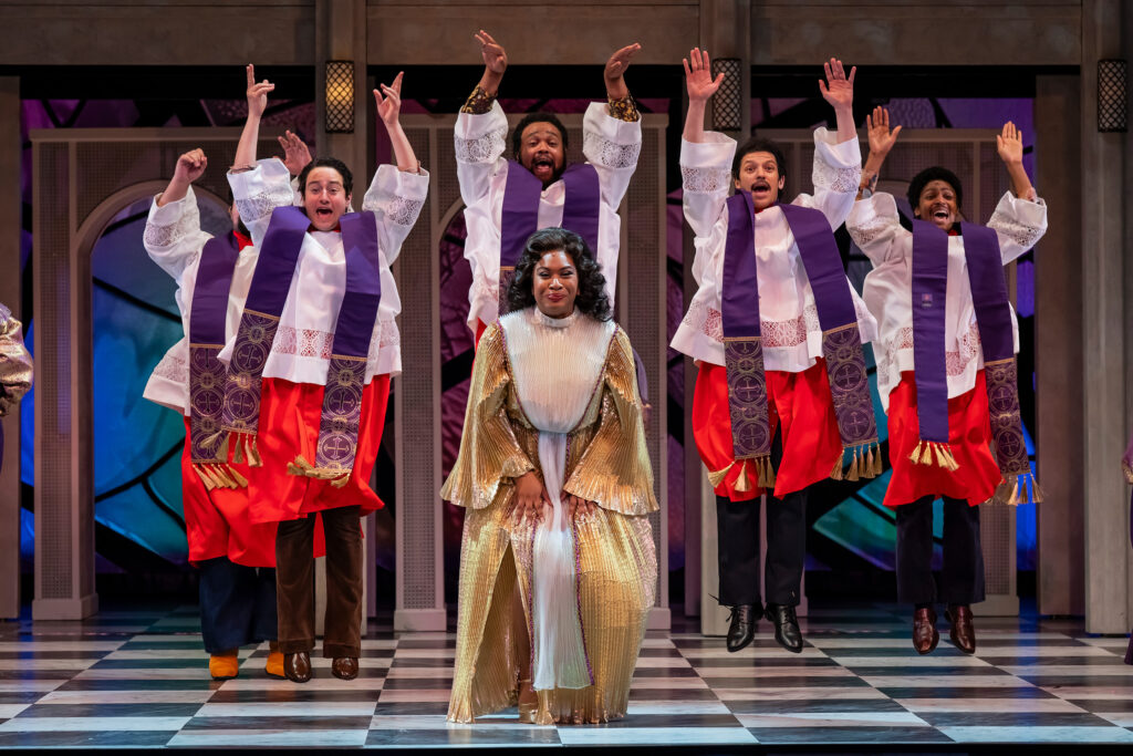A Black woman in a gold and white gown dances in front of a group of dancing men all dressed in Catholic priest attire.