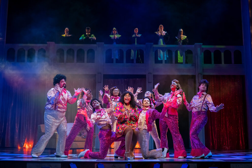 A Black woman in a colorful 1970s-era dress sings while sitting on the knees of two male performers. Behind them is a group of performers, all dressed in shimmery bright pink and white 1970s-era costumes. On an elevated platform at the rear of the stage, nuns in habits look on at the dancers in prayer.