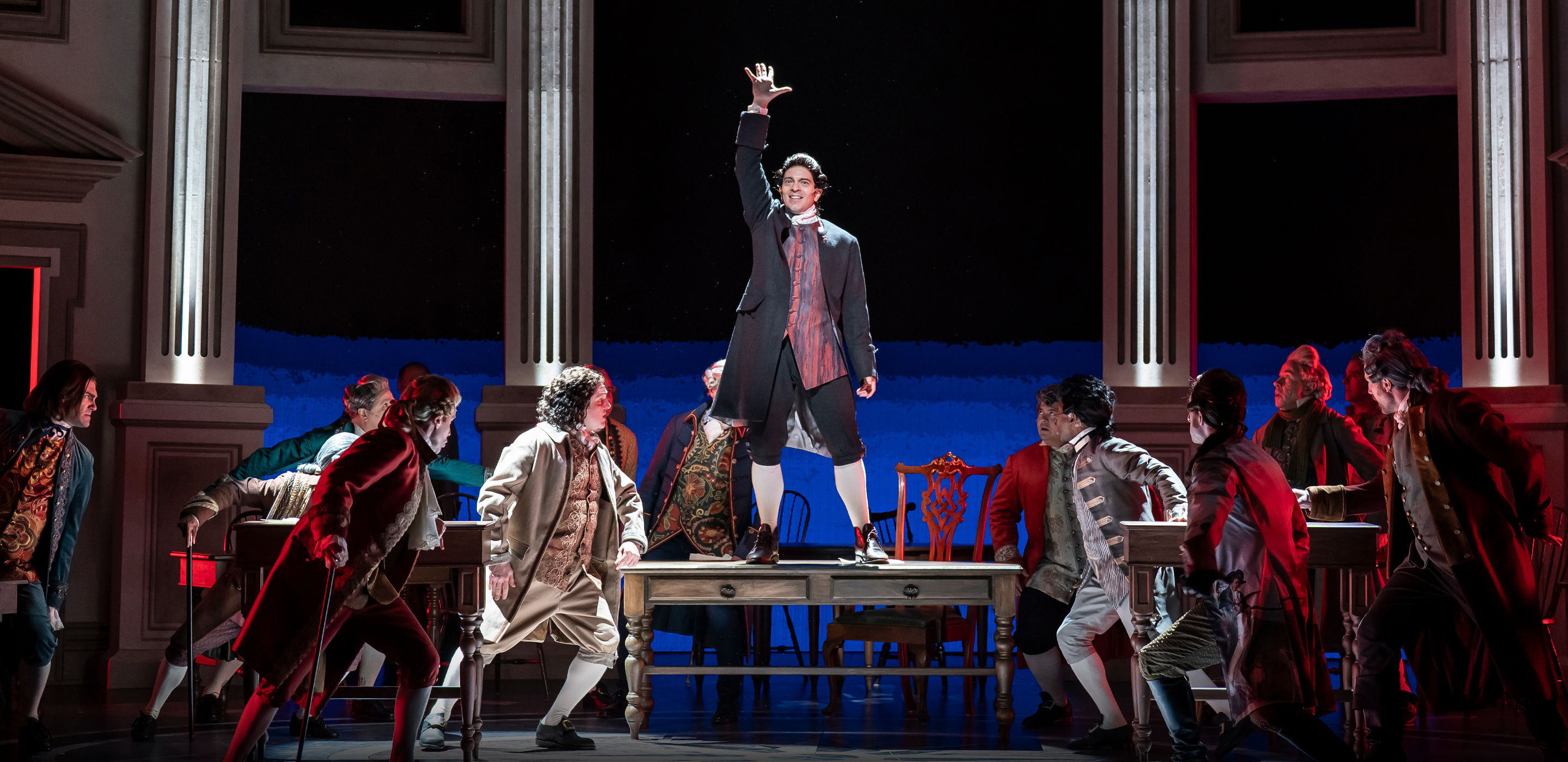 A man in an 18th‑century coat stands triumphantly on top of a table with one arm raised, surrounded by other actors in period costumes who look up toward him. The dramatic lighting and grand colonial‑style set heighten the intensity of the scene.