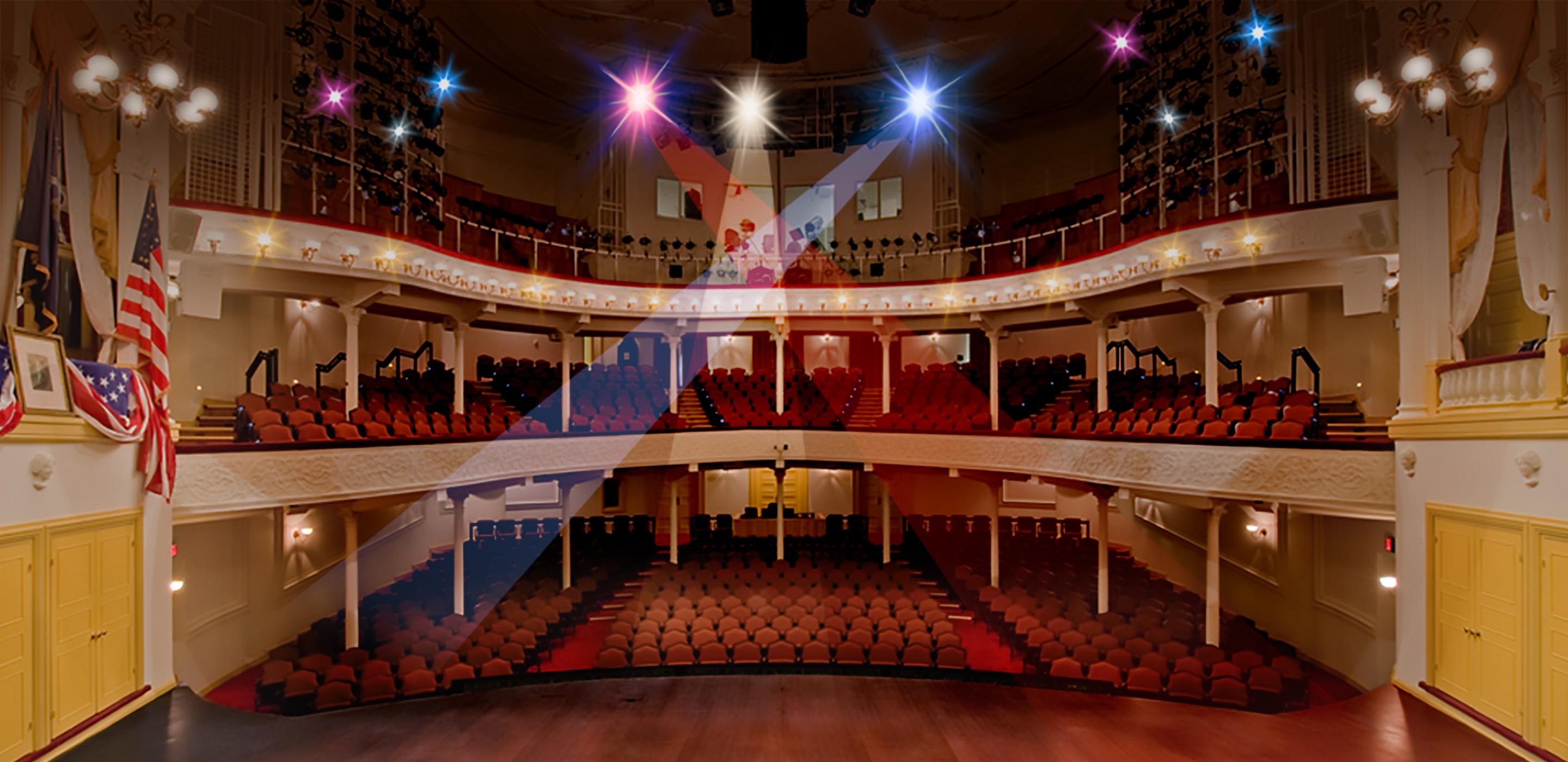 The view from the Ford’s Theatre stage looking out to the audience. To the left of the stage is the President Box with an American flag, a framed picture of George Washington and American flag bunting draped over the box. To the right is another box with yellow and white curtains. In the center of the stage is a wooden desk. The view includes two levels of seating and rows of lighting equipment on the third level.