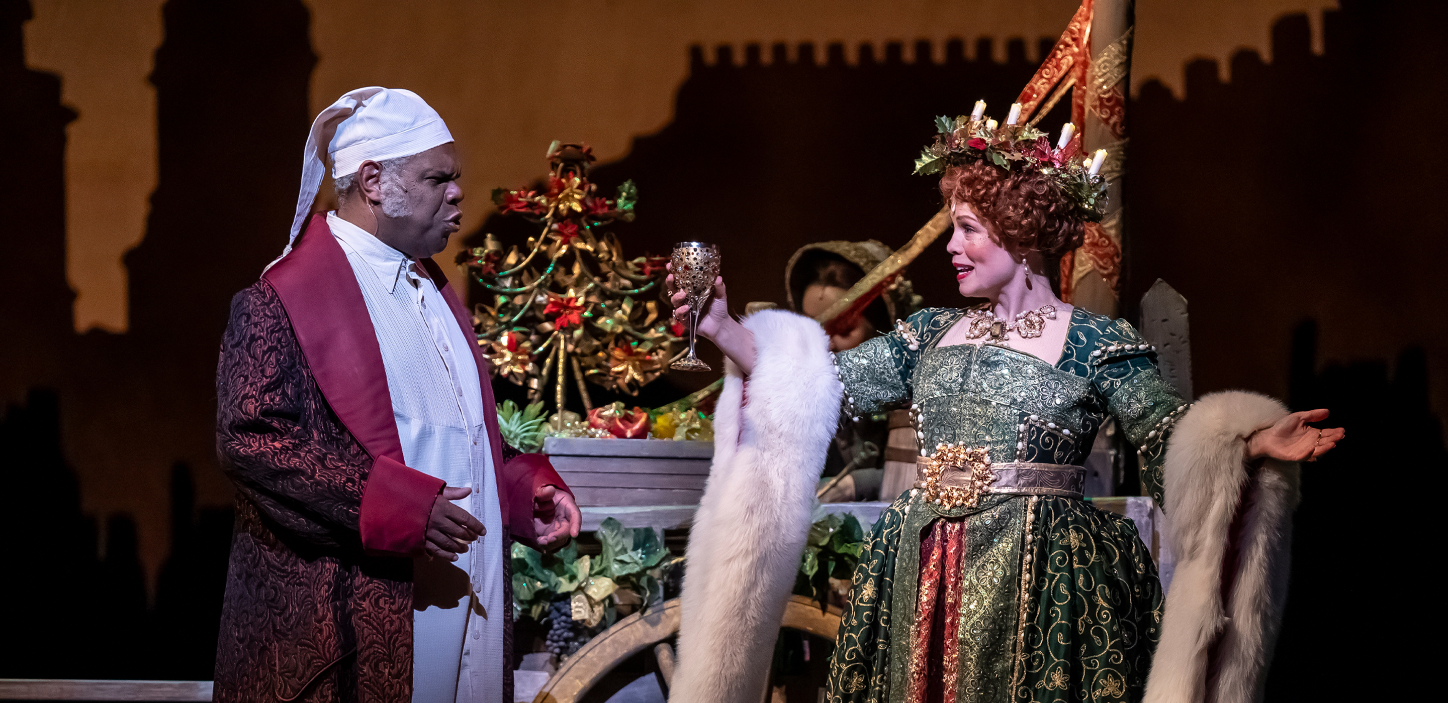 A woman and man stand next to a decorative cart: a man in a long, dark red dressing robe and white nightcap speaks to a woman in a green, gold and red gown draped with a white fur stole.