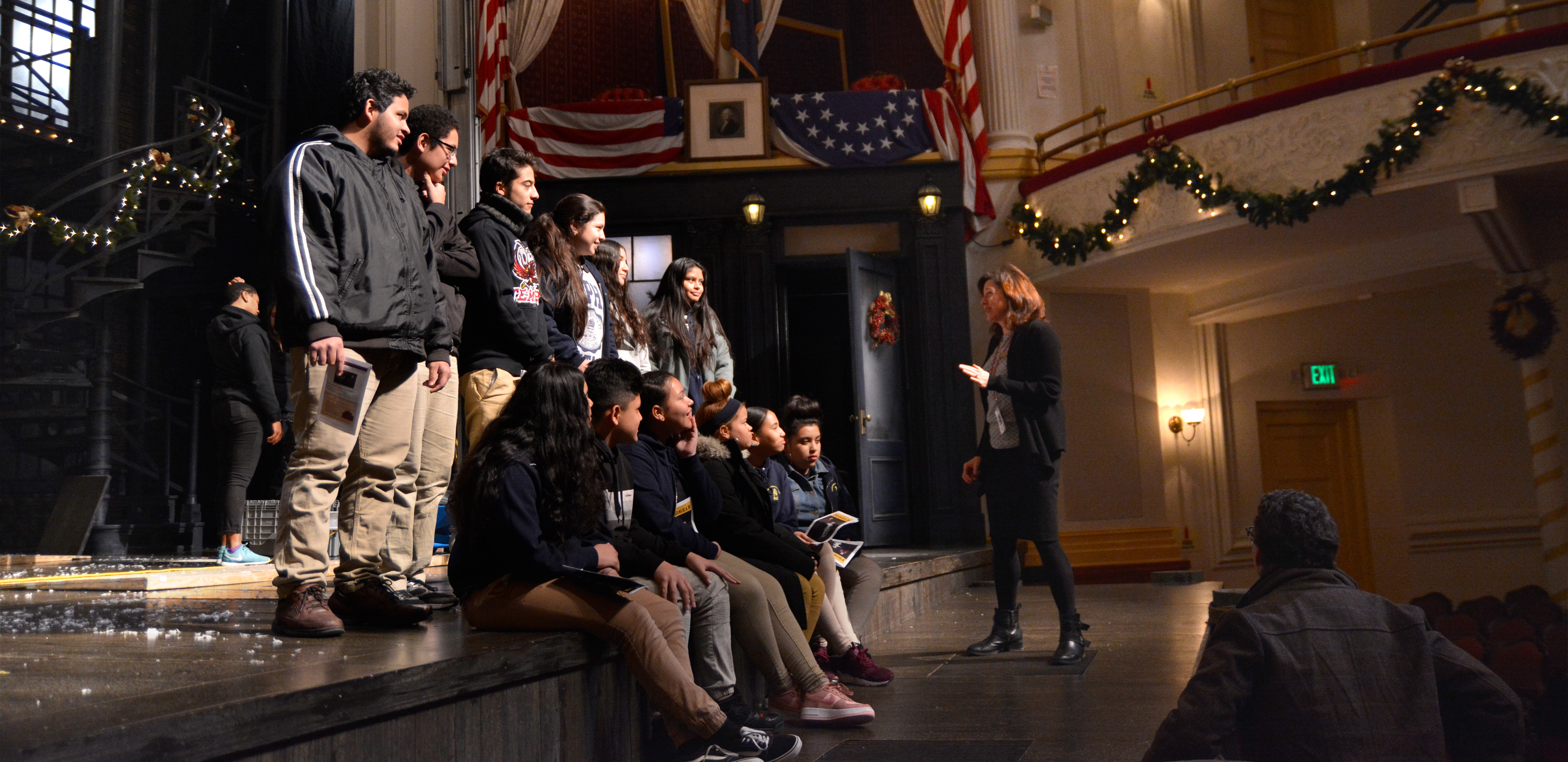A group of students, half sitting and half standing, wait on a stage in a theatre while a teacher speaks to them.