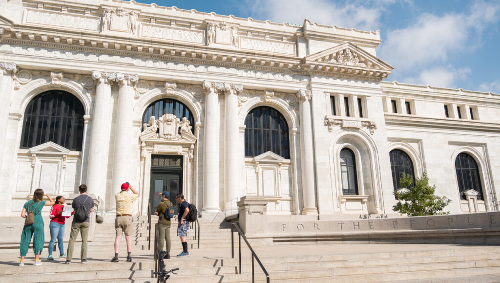 A group of tourists stand in front of a large marble building and take pictures.