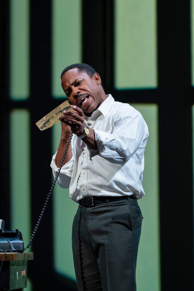 A Black man, wearing a white button-up shirt and dark gray pants, holds a telephone receiver to his ear and holds a slip of paper in his other hand, and is speaking into the mouthpiece.