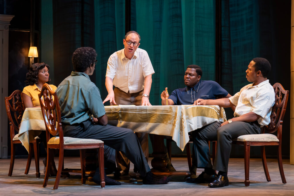 A white man in a white shirt and tan pants stands at the head of the table, leaning forward and speaking with an open mouth. Two Black men are seated to his right, and a Black man and a Black woman are seated to his left. The man closest to the camera on the right is pointing at a piece of paper on the table.