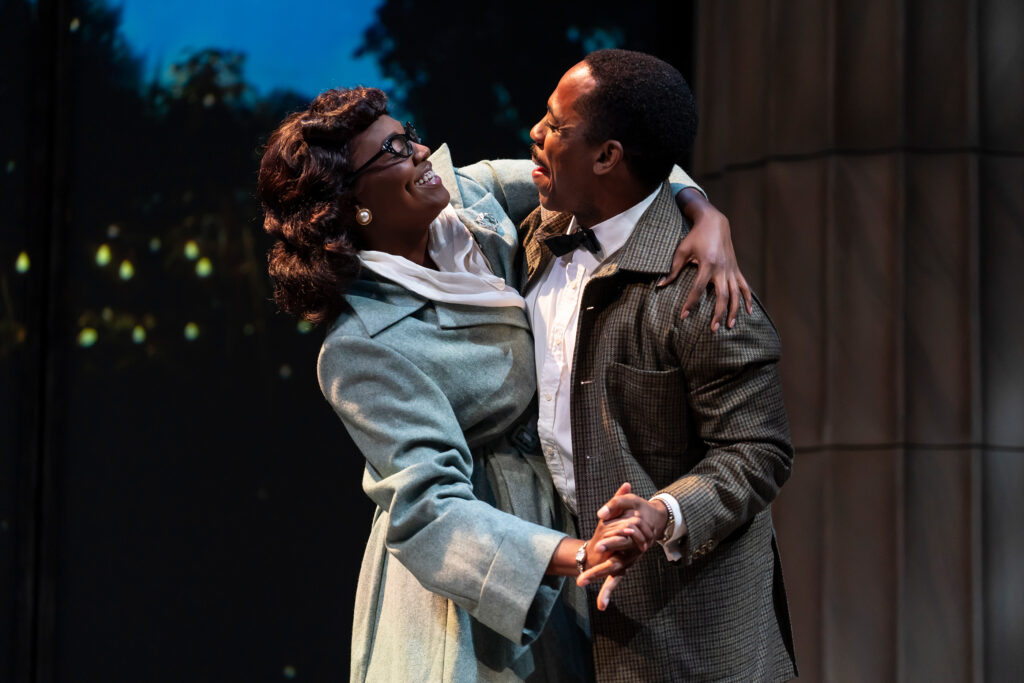A Black man, dressed in a checkered jacket, white shirt and bow tie, and a Black woman, wearing glasses, pearl earrings and a light blue coat, smile at each other while dancing.