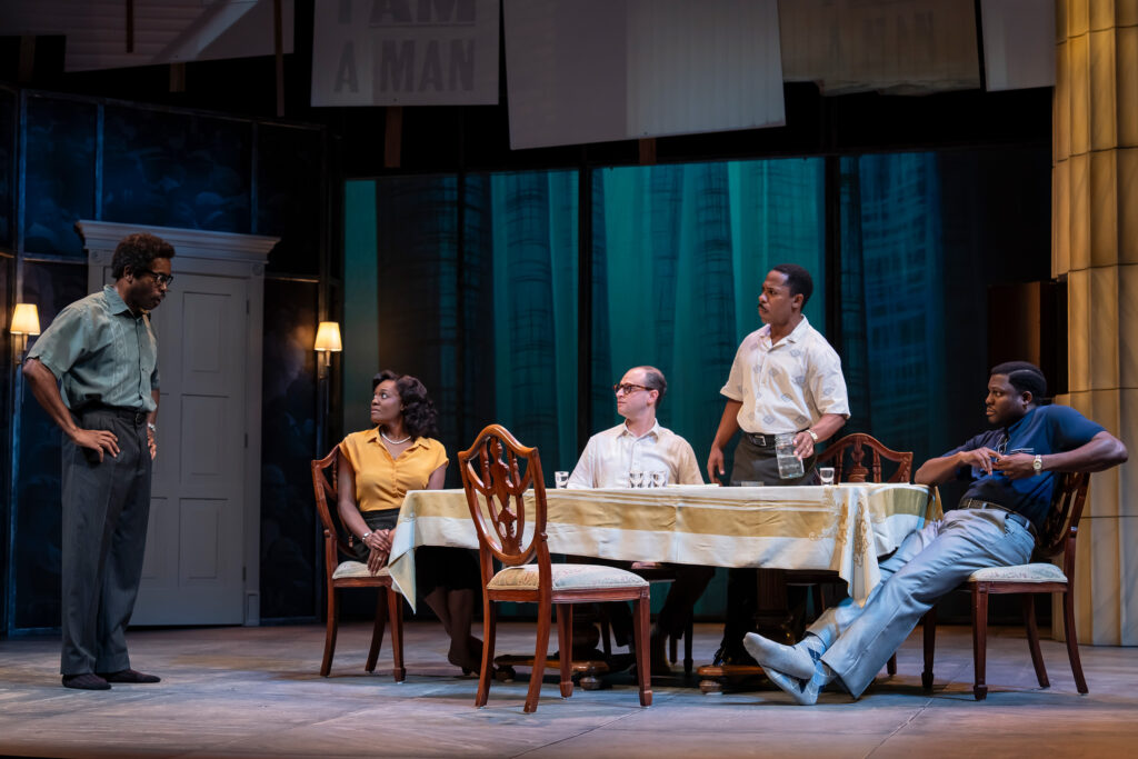 A Black man in a green short-sleeved shirt and gray pants stands on the left, looking toward the group at the table with his hands on his hips. At a table with a white and yellow tablecloth, are four people: a Black woman in a yellow blouse and black skirt, a white man in a white short-sleeved shirt, a Black man in a white short-sleeved shirt and dark pants who is standing and holding a glass, and a Black man in a dark blue polo shirt and gray pants who is leaning back in his chair.