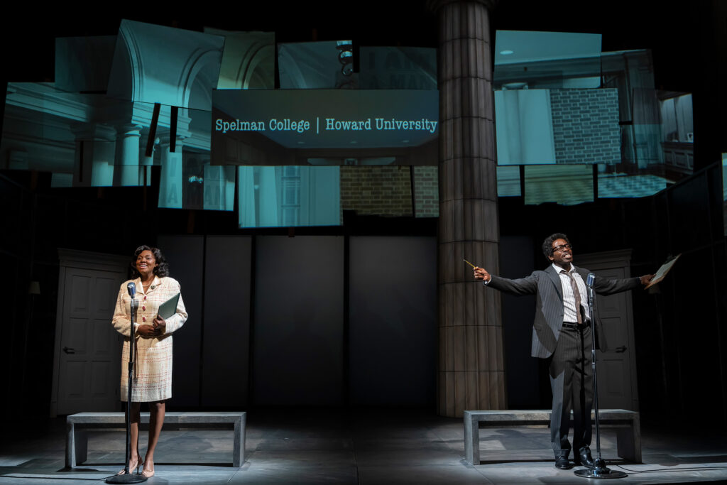 A Black woman stands on the left, wearing a light-colored tweed dress, behind a microphone stand, holding a book and smiling. A Black man stands on the right, wearing a dark suit with a white shirt and a dark tie, holding a sheet of paper. Large screens above them display the words "Spelman College | Howard University."