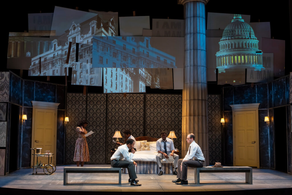 A Black woman wearing a patterned dress stands on the far left next to a silver serving cart with drinks. Two men, a Black man and a white man, both in white shirts and dark pants, sit facing each other on a bench at the foot of a bed. A third man, also a Black man in a white shirt and dark pants, sits on the bed between them.