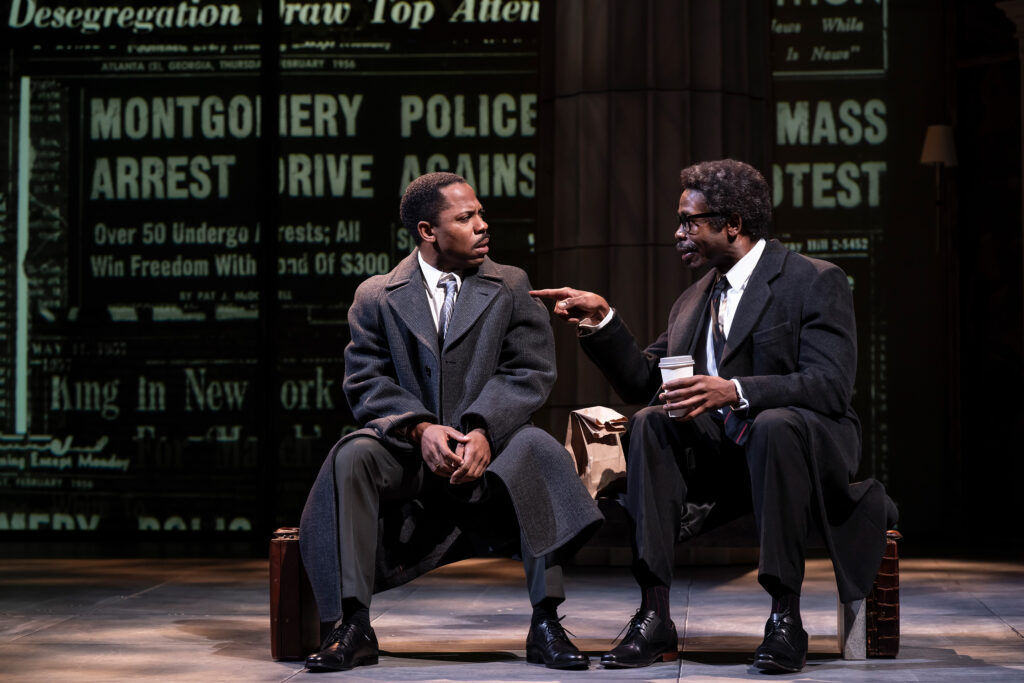 A Black man, dressed in a gray overcoat and tie, sits on a bench with a suitcase at his feet, looking toward another Black man in a dark suit and overcoat who gestures while holding a coffee cup. A newspaper headline about the Montgomery bus boycott is projected in the background.