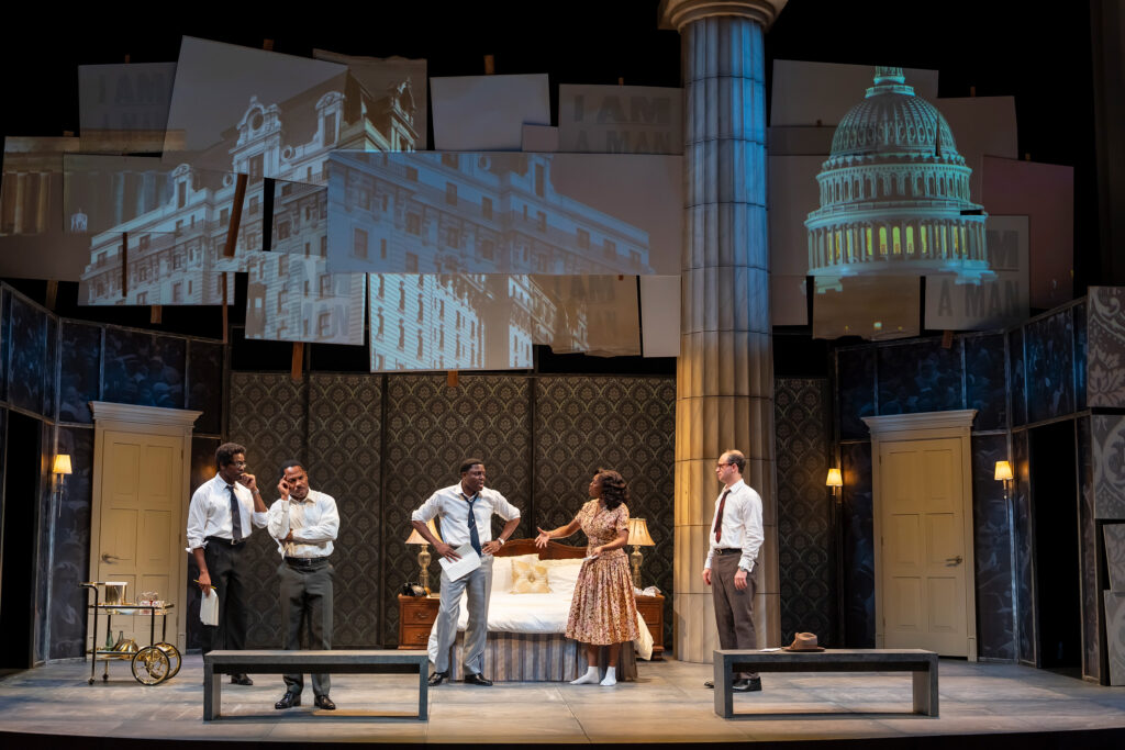 A Black man in a white shirt and dark pants stands on the left, holding a piece of paper. Next to him, a Black man in a white shirt and dark pants holds his head. A Black man in a white shirt and light gray pants stands with a Black woman in a patterned dress beside a bed in the center. At the far right, a white man in a white shirt and dark pants, who is looking at the others.