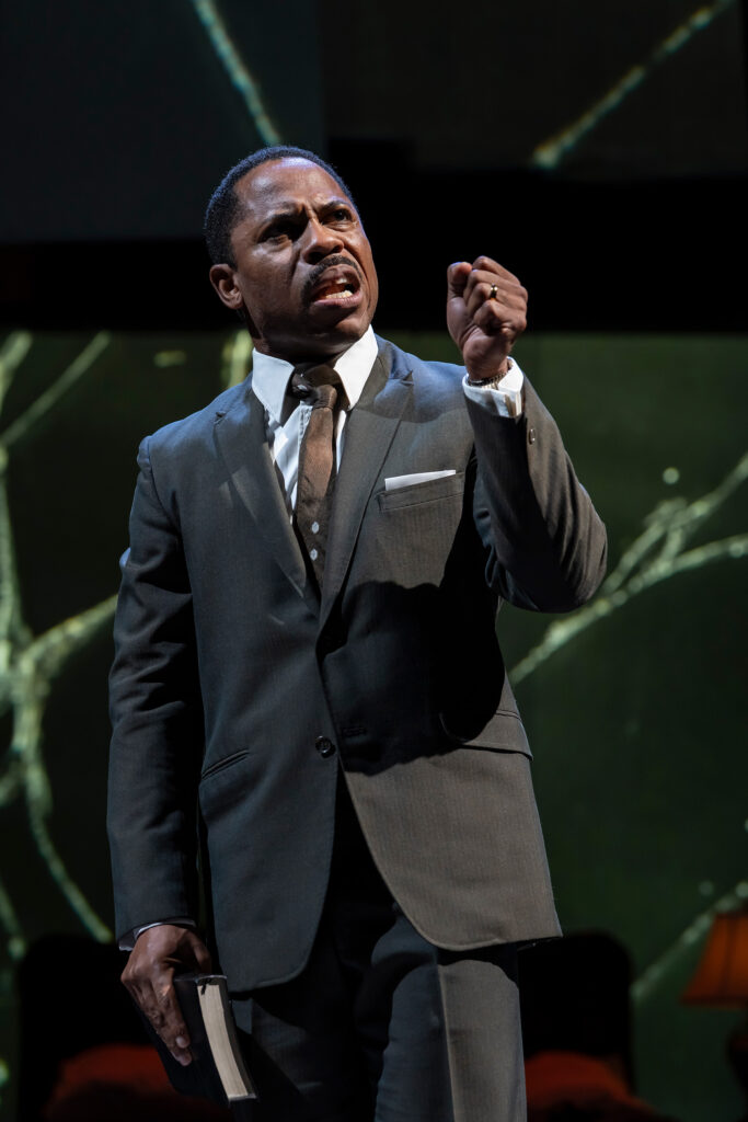 A Black man in a dark gray suit with a white shirt and a brown tie stands with his mouth open, speaking passionately. He clenches his right hand into a fist, raising it slightly.