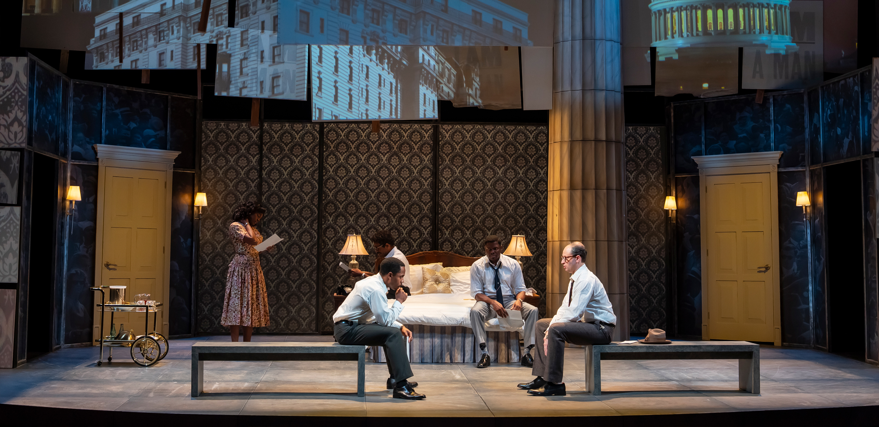 A Black woman wearing a patterned dress stands on the far left next to a silver serving cart with drinks. Two men, a Black man and a white man, both in white shirts and dark pants, sit facing each other on a bench at the foot of a bed. A third man, also a Black man in a white shirt and dark pants, sits on the bed between them.