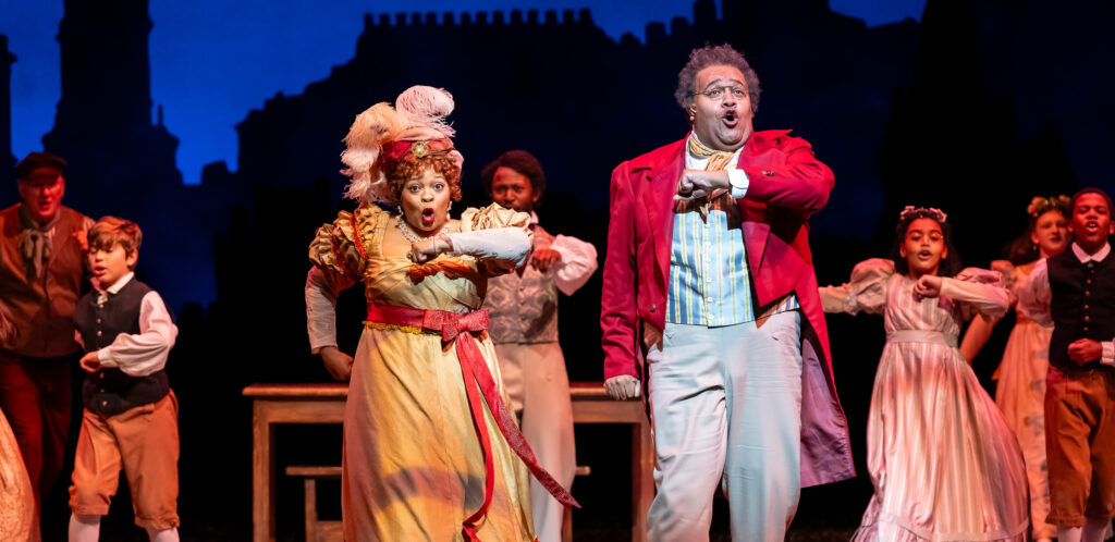 A woman and a man dressed in red and gold elegant Victorian-era party clothes dance at a holiday party.