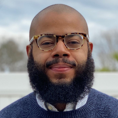 Headshot of a Black man with a beard and glasses wearing a blue sweater.