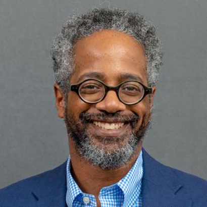 Headshot of a Black man with grey hair, a beard and glasses wearing a blue blazer and blue shirt.