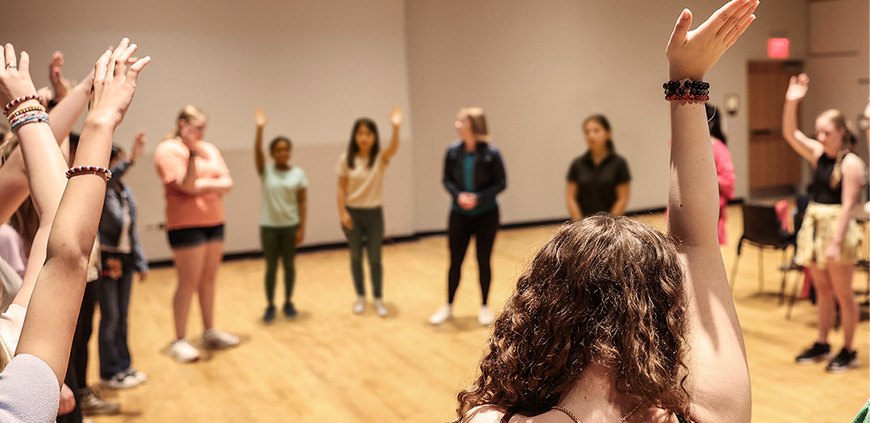 A group of girls stand in a circle in an auditorium, many with their hands raised.