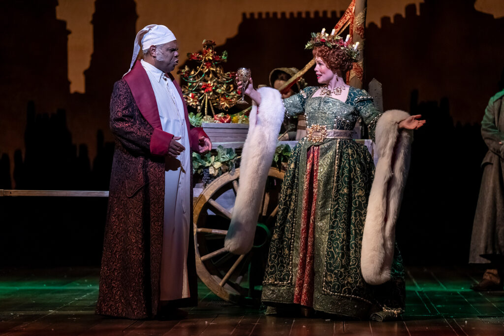 A woman and man stand next to a decorative cart: a man in a long, dark red dressing robe and white nightcap speaks to a woman in a green, gold and red gown draped with a white fur stole.