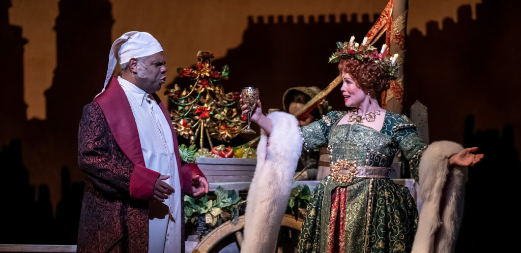 A woman and man stand next to a decorative cart: a man in a long, dark red dressing robe and white nightcap speaks to a woman in a green, gold and red gown draped with a white fur stole.
