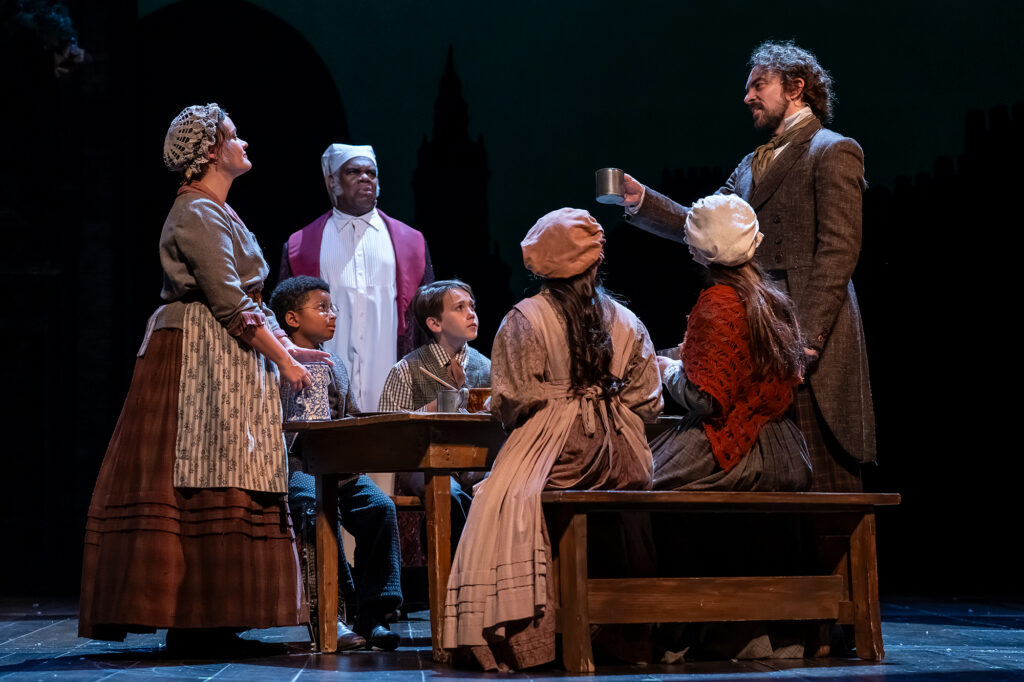 A woman and man stand with a family seated at a wooden table on stage: a man in a dark brown coat and vest holds a mug toward a woman in a bonnet seated on the right, while a woman in an apron and bonnet stands next to a boy and a man in a long white nightshirt stands in the background.