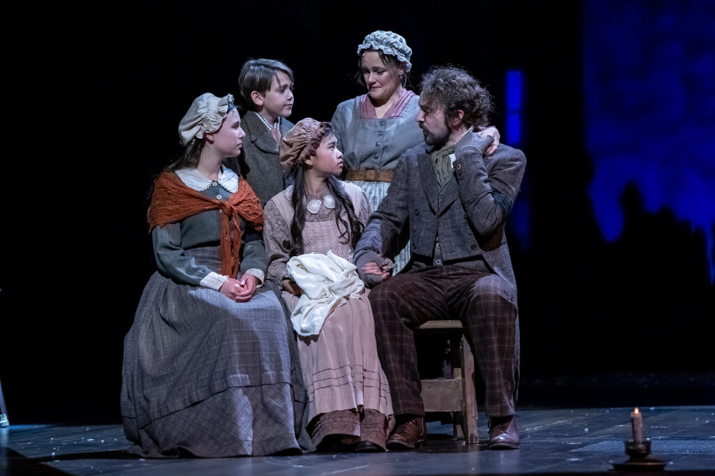 A family of five is together on a dark stage; a man in a textured coat sits holding hands with a young woman in a bonnet, surrounded by two girls and a boy, all dressed in period clothing.