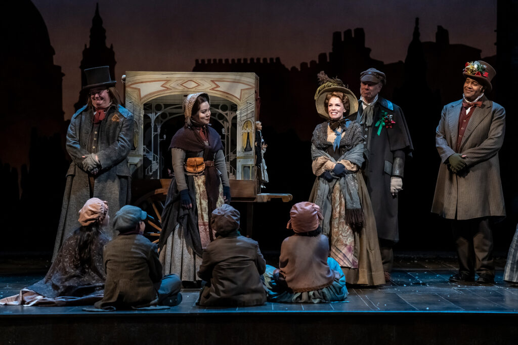 A group of people in Victorian-era costumes stand smiling before a painted street-cart set piece, while several children in period clothing sit on the ground facing them, creating a lively and cheerful scene from a holiday production.