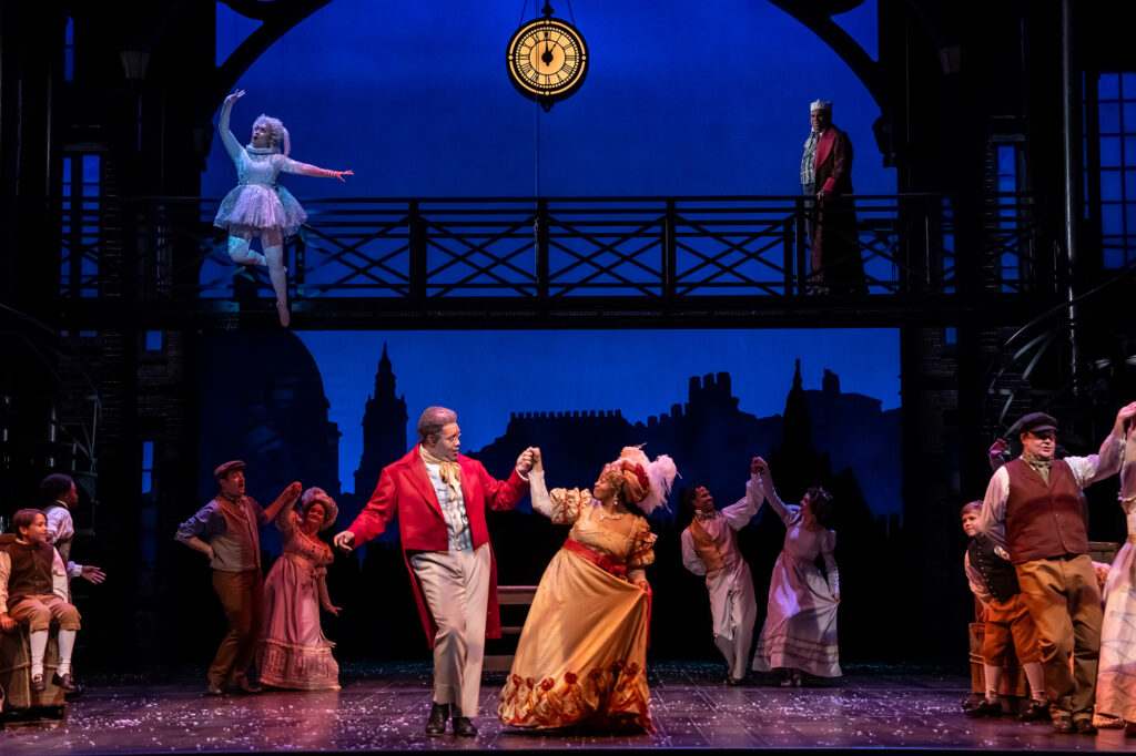 A man in a vibrant red Victorian jacket dances with a woman in a shimmering gold dress on a stage surrounded by other period-costumed performers, while above them, a figure in a dark costume watches from a bridge, and a person in an icy-white gown floats mid-air.