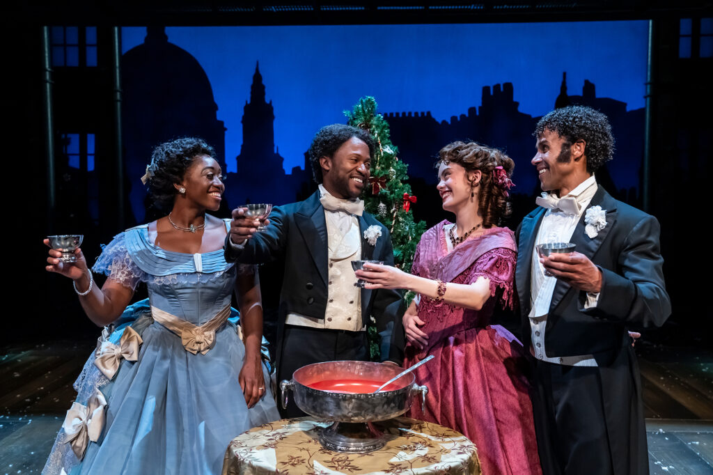 A festive group of two couples in ornate Victorian-style attire stands around a small table with a silver bowl of red punch, raising silver cups for a toast, all smiling brightly against a deep blue backdrop of silhouetted buildings and a Christmas tree.