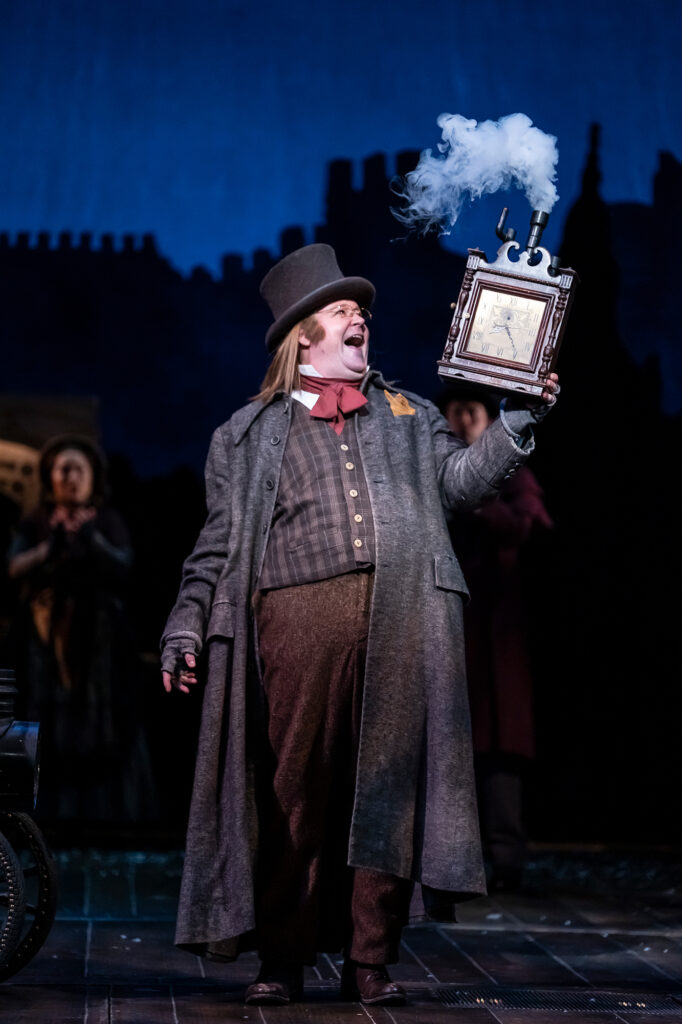 A man, wearing Victorian-style clothing, including a long gray coat, plaid vest, red cravat, and tall top hat, smiles broadly while holding up a wooden, clock-like prop that releases a plume of white smoke. 