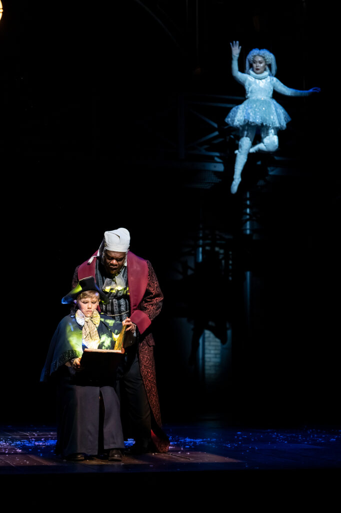 A man dressed in ornate Victorian-style nightclothes leans over a child in period attire who is seated and gazing into a glowing book, while above them a person in a shimmering, icy-white costume floats midair with one arm raised.
