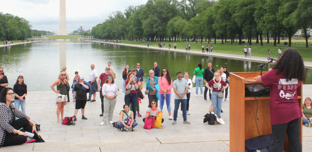 A high school student stands at a podium speaking to a group of attentive listeners. The crowd in gathered in front of the Reflecting Pool with the Washington Monument in the background