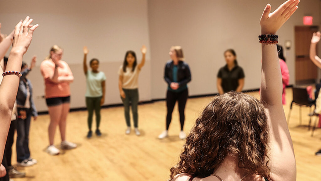 A group of girls stand in a circle in an auditorium, many with their hands raised.
