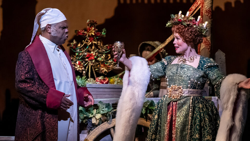 A woman and man stand next to a decorative cart: a man in a long, dark red dressing robe and white nightcap speaks to a woman in a green, gold and red gown draped with a white fur stole.