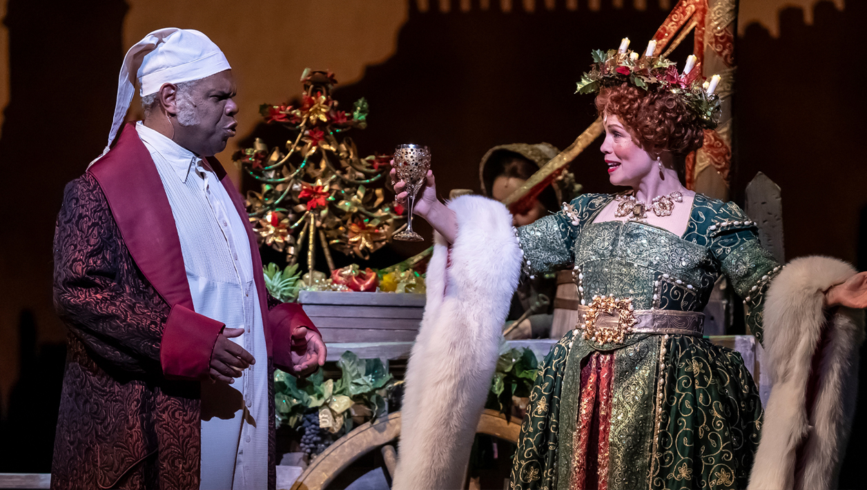 A woman and man stand next to a decorative cart: a man in a long, dark red dressing robe and white nightcap speaks to a woman in a green, gold and red gown draped with a white fur stole.