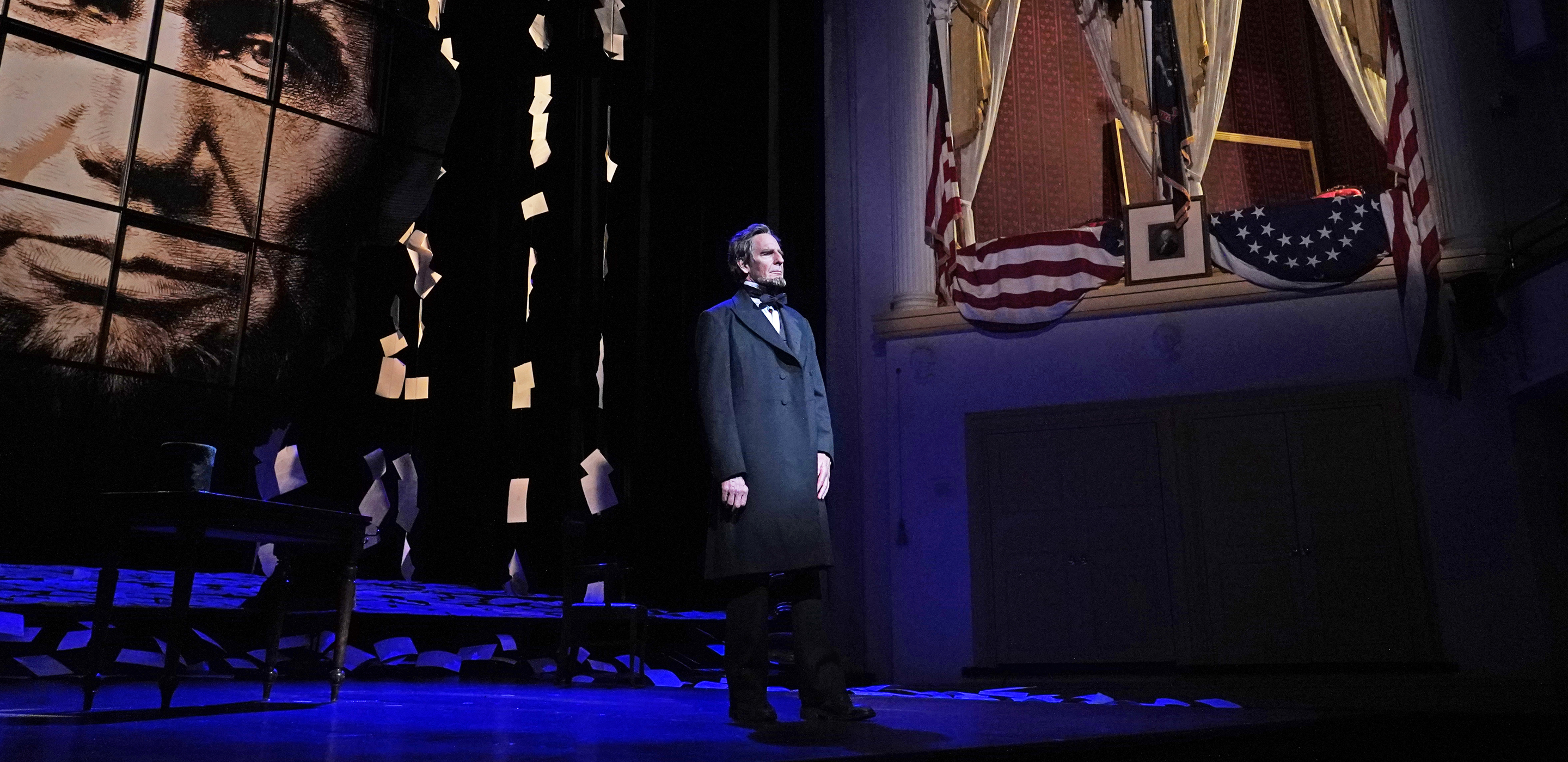 Abraham Lincoln stands on a stage with a large picture of his face to his left and a lit seating box to his right.