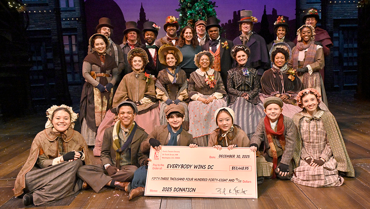 A group of adults and children in victorian dress stand or kneel behind a large check made out to Everyone Wins DC.